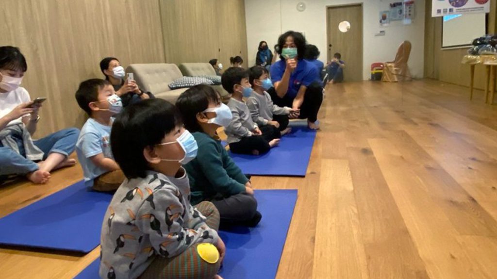 忍者小學堂的小朋友整齊坐在藍色墊子上，在木質地板教室中專注聆聽前方教練的課程指引。畫面捕捉孩子在團體活動中的靜心觀察與自律表現，展現 Oovi 正念引導教學的核心價值。