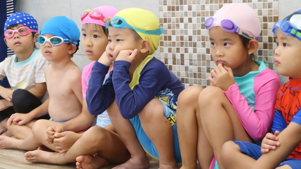 一群穿著色彩繽紛泳裝的孩子戴著泳帽與泳鏡，在游泳池邊排排坐，全神貫注地盯著教練示範。特寫畫面呈現小朋友認真觀察、等待指令的專注神情，這是水中忍者任務開始前的心理建設與專注力培養。
