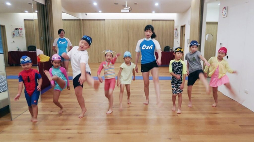 Oovi 忍者小學堂活動紀錄。穿著藍色 Oovi 制服的教練帶領一群穿著泳裝與運動服的孩子在木地板教室進行踢腿動作練習，透過趣味互動開發兒童大肌肉群、平衡感與身體協調能力。