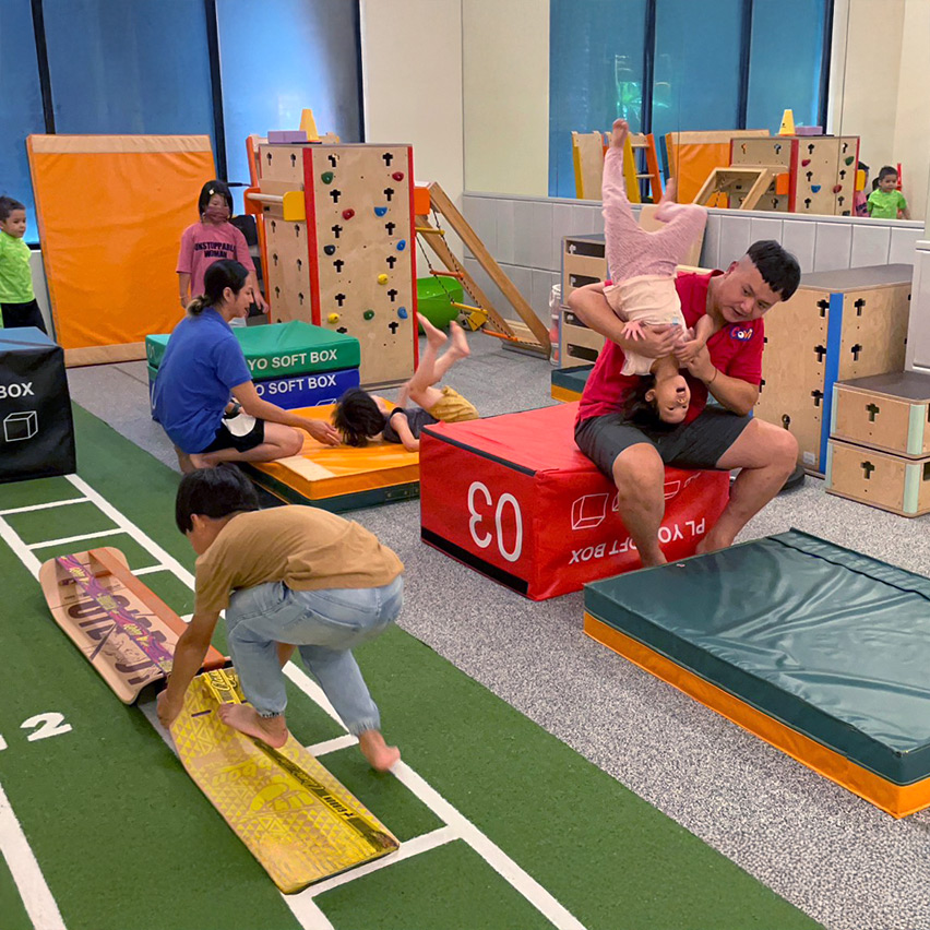 呈現 Oovi 室內教室的多元配置，包含攀岩牆、平衡板與軟墊區域，專業老師協助孩子練習倒立與翻滾，提供豐富的本體覺刺激。