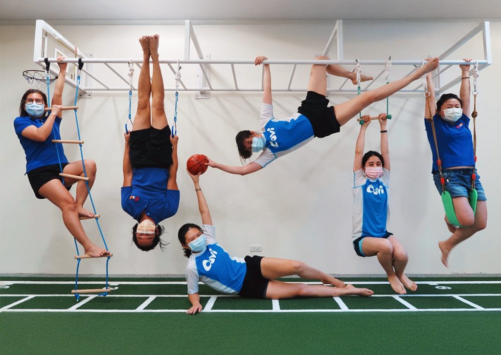 Oovi 老師們在專業陸上體能教室（FMS 設施）展現俏皮且專業的一面。有的老師輕鬆懸掛在攀爬架上，有的老師傳遞籃球，展現出卓越的身體素質與幽默感。團隊身穿 Oovi 專業制服，呈現出一個愛玩、敢挑戰且極具感染力的工作氛圍，是吸引熱血教育人才加入的最佳職涯寫照。