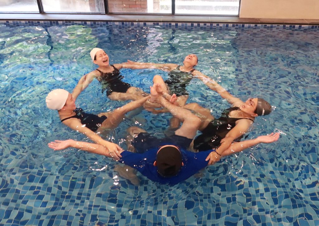 Oovi 師資團隊在室內溫水泳池中圍成一個和諧的圓圈，老師們彼此連結、向後仰漂並露出燦爛且真誠的笑容。這張照片展現了 Oovi 教師充滿活力、溫暖且熱愛團隊合作的人格特質，完美詮釋了「先照顧好自己，才能陪伴孩子」的教學溫度。