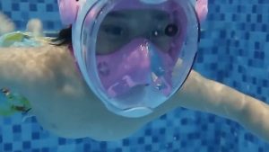 一名參與 Oovi 親水成長探索營隊的小朋友戴著粉色全臉式浮潛面罩，在充滿清澈藍色瓷磚的游泳池中潛水游動。畫面呈現孩子在專業教練引導下，透過裝備輔助克服對水的恐懼，建立卓越的水中自信心與肢體協調力。